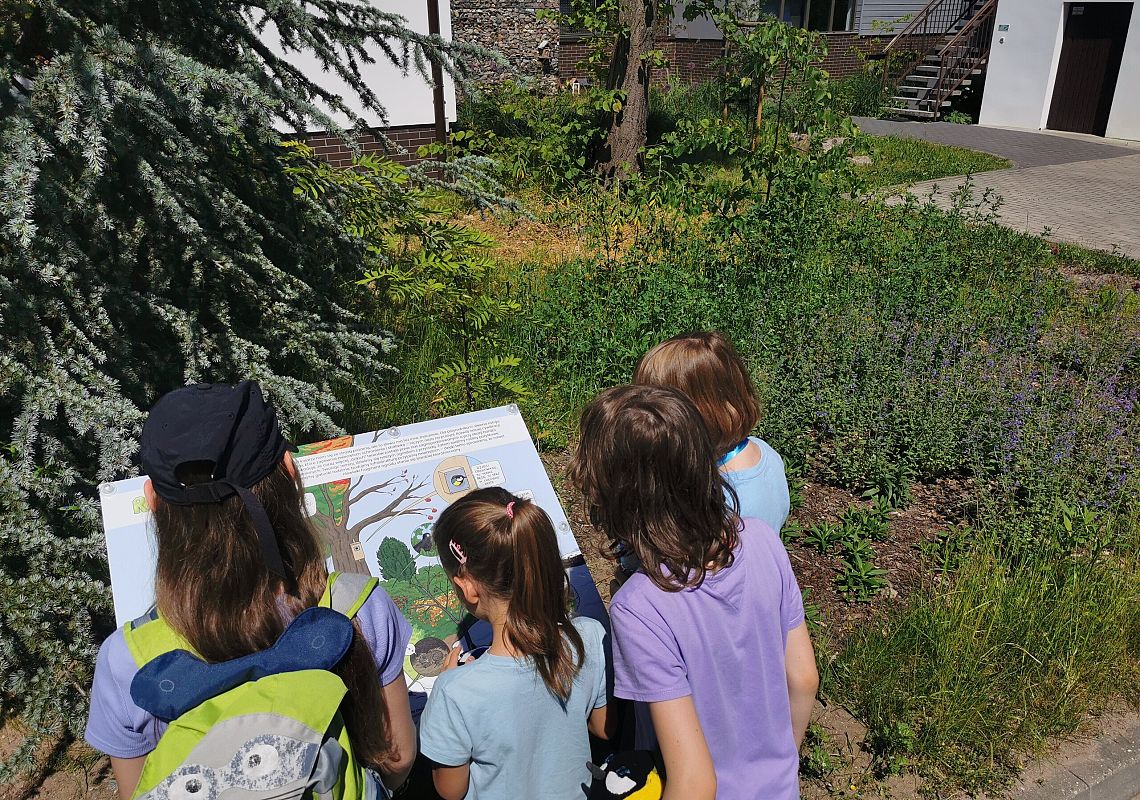 Przy remizie, fot. JK Czórka dzieci stoi przed tablicą edukacyjną w stylu komiksowym, w tle ogród i budynek