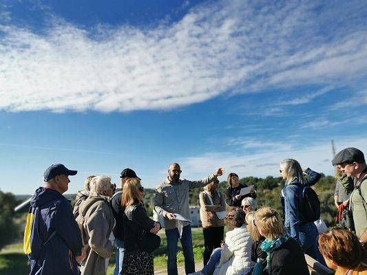 Mężczyzna ze starymi maami w ręku wśród grupy słuchaczy. W odali zieleń, niebieskie niebo.