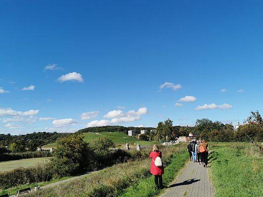 Grupa dorosłych idzie po chodniku, teren zielony, po lewej stronie woda, na wprost pagórek porośnięty trawą.