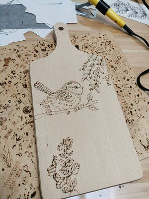 drewniana deska kuchenna z wypalonym obrazkiem ptaka i roślin. w tle kolba od pirografu, kartki i inne deseczki.
