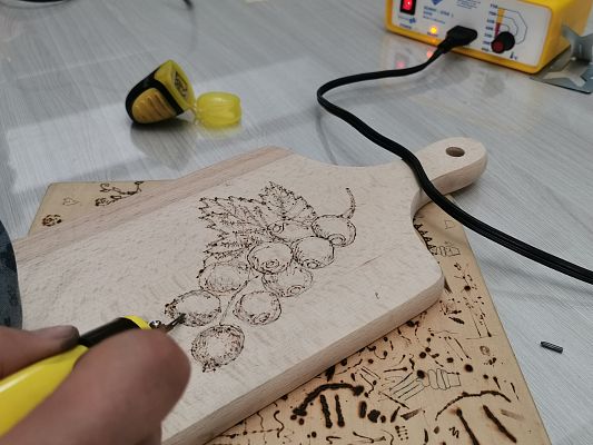 Zbliżenie na dłoń, która trzyma kolbę od pirografu i wypala owoce i liście na drewnianej desce.