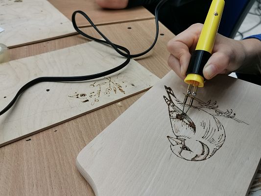 Zbliżenie na dłoń, która trzyma kolbę od pirografu i wypala wizerunek ptaka na drewnianej desce.