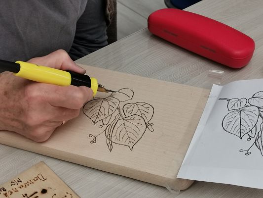 Zbliżenie na dłoń, która trzyma kolbę od pirografu i wypala liście lipy na drewnianej desce.