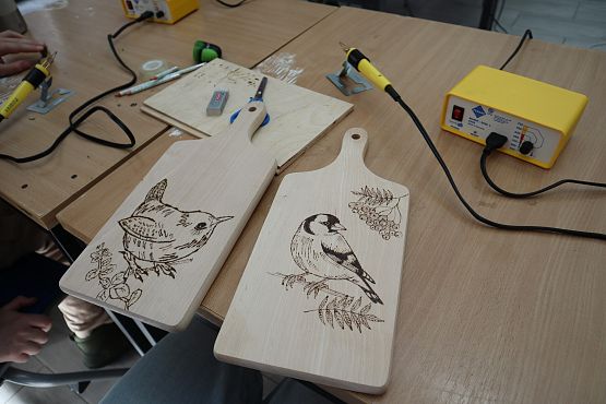 dwie drewniane deski kuchenne ozdobione wypalonymi pirografem rysunkami, na obu wizerunek ptaków. Obok leżą pirografy.