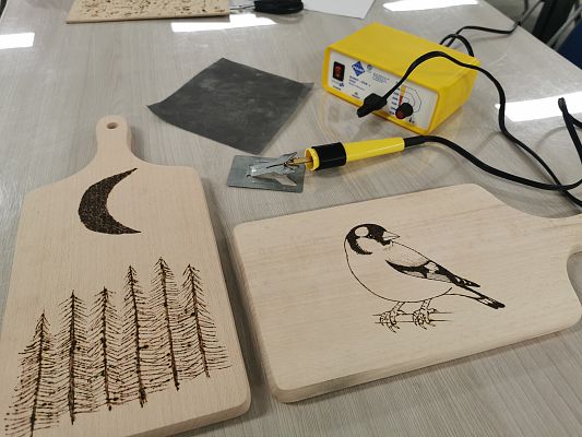 dwie drewniane deski kuchenne ozdobione wypalonymi pirografem rysunkami, na jednej z nich drzewa i księżyc a na drugiej ptak. Obok leży pirograf.