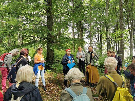 Grupa osób stoi w kręgu w lesie, słucha opowieści kobity z różowymi krótkimi włosami.