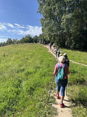 Grupa osób idzie ścieżką, jedna za drugą, pod górkę, Po prawej stronie las, po lewej trawa. Pierwsza osoba idzie z kilkami.