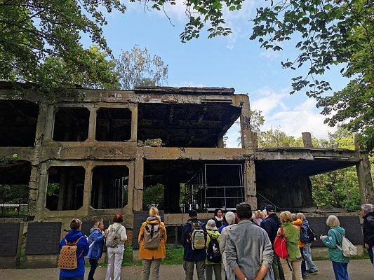 Ruiny budynku zniszcoznego podczas wojny. Przed budynkiem grupa ludzi, odwrócona plecami, słucha przewodniczki.