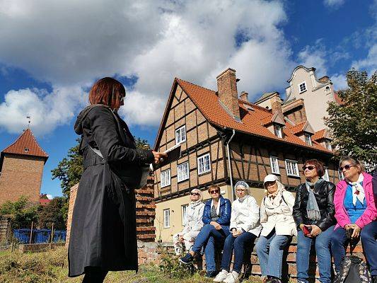 Sześc kobiet siedzi na mursku z cegły. Przed nimi stoi kobieta ubrana na czarno. Za nimy budynek z cegły i drewnainych bali.