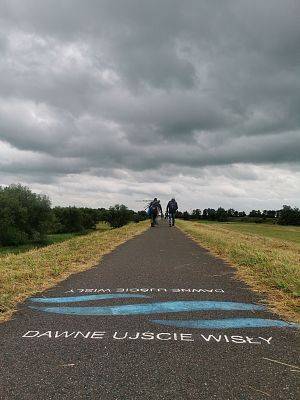 Asfalowa ścieżka na wale, w oddali idzie grupa osób, jedna osoba niesie statyw. Na ścieżce napis: dawne ujście Wisły i trzy niebieskie fale.