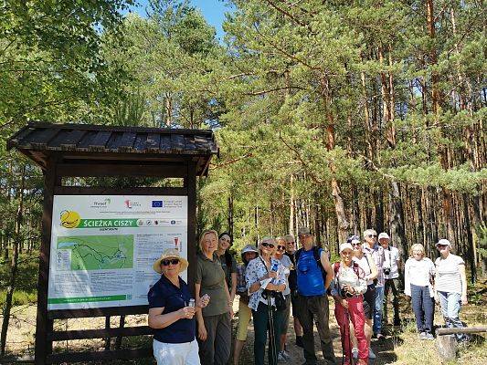 Grupa osób stoi w lesie, przy drewanianej tablicy edukacyjno-informacyjnej o ścieżce ciszy. Na tablicy mapa, tekst i logotypy.