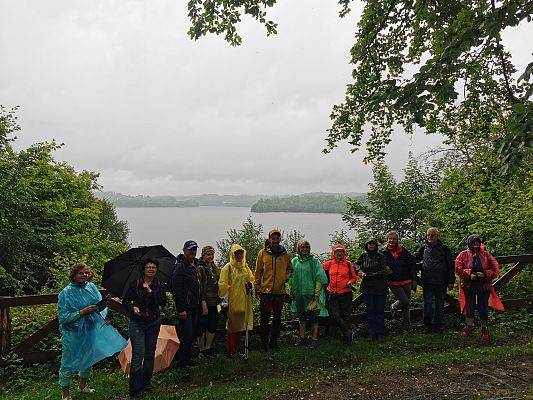 Grupa osób z płaszczach przeciwdeszczowych i z parasolami, stoi na punkcie widokowym nad jeziorem.