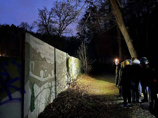 grupa ludzi na ścieżce w lesie o zmorku, świecą latarkami. stoją przy betonowym płocie na którym jest namalowany mural i jest tam tablica informacyjna