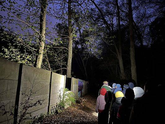 grupa ludzi na ścieżce w lesie o zmroku, świecą latarkami. stoją przy betonowym płocie