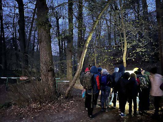 grupa ludzi o zmroku, stoją przy szlabanie prowadzącym do lasu, świecą latarkami